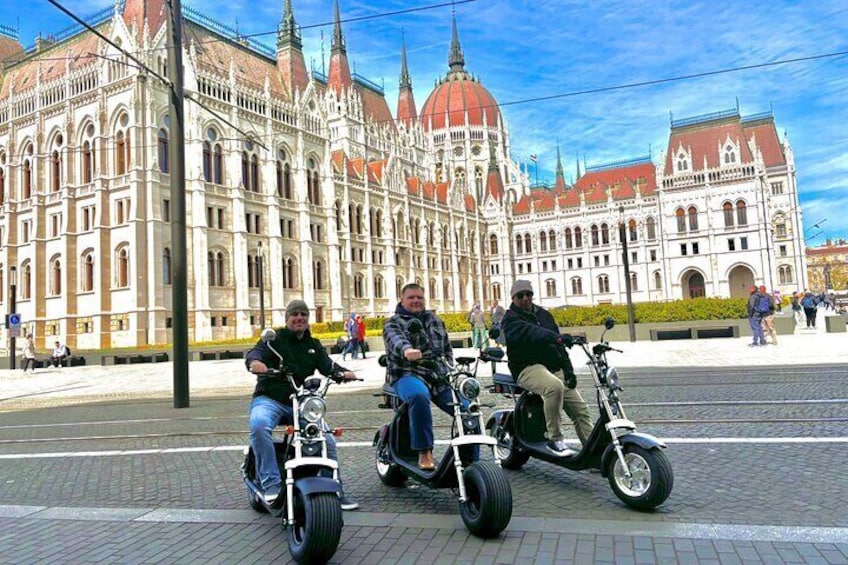 Three people riding electric scooters in front of a historic building with ornate architecture. 1