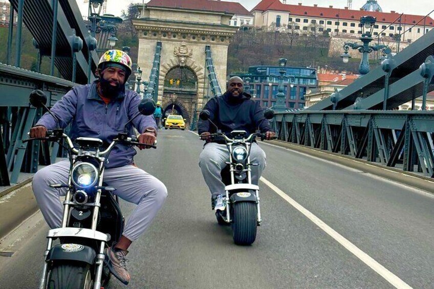 Two people riding motorcycles on a bridge with a historic arch in the background. 4