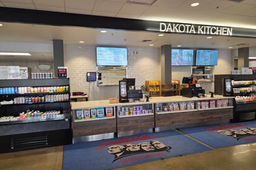 A food court with a sign reading 'Dakota Kitchen,' a digital menu board, and a variety of food and drink options. 2