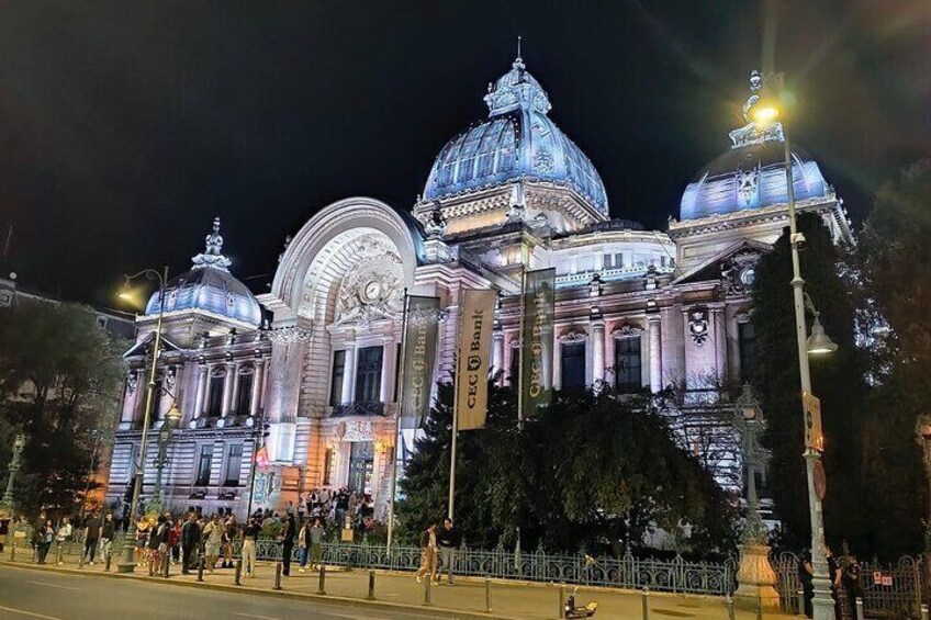 A grand, illuminated building with domed structures and columns, surrounded by a fence and people. 1