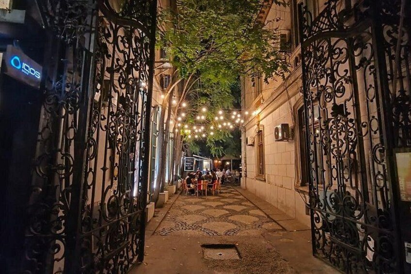 A narrow alley with wrought iron gates, string lights, and a few people sitting outside a building. 3