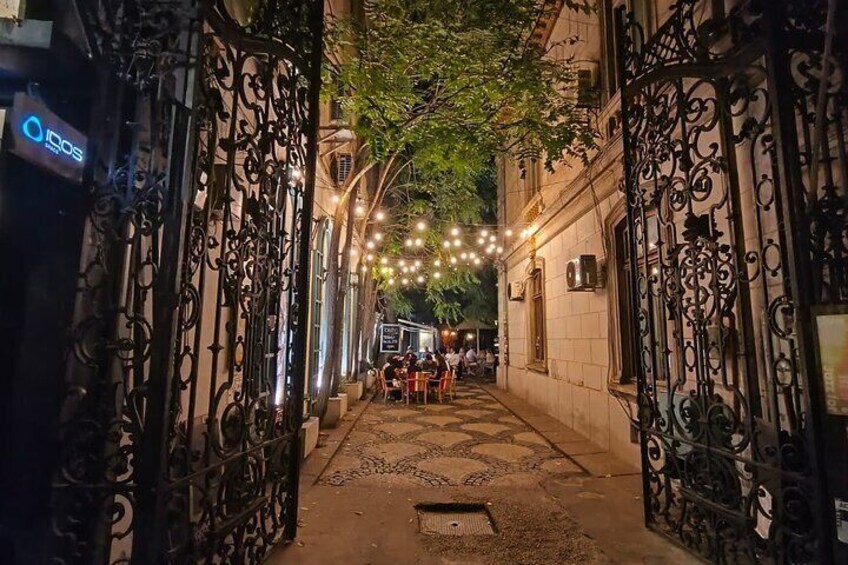 A narrow alley with ornate wrought iron gates, string lights, and a few people sitting at tables. 3