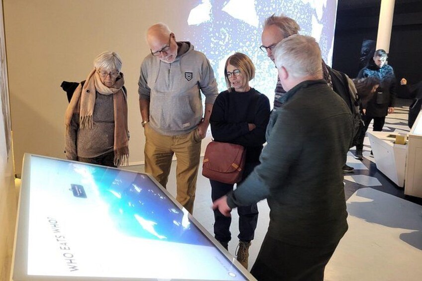 Eine Gruppe von Menschen in einem modernen Innenraum, möglicherweise ein Museum oder eine Ausstellung, mit einer großen digitalen Anzeige im Vordergrund. 5