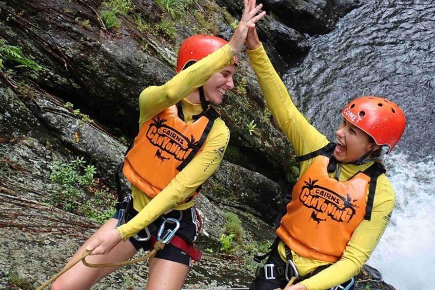 Two people wearing helmets and life jackets, high-fiving near a rocky waterfall. 3