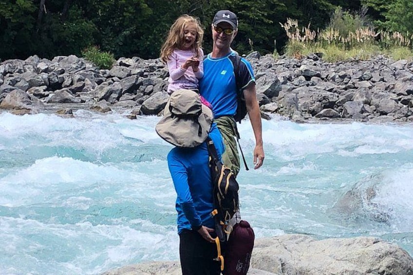 A person standing on rocks by a flowing river, carrying a child on their shoulders. 2