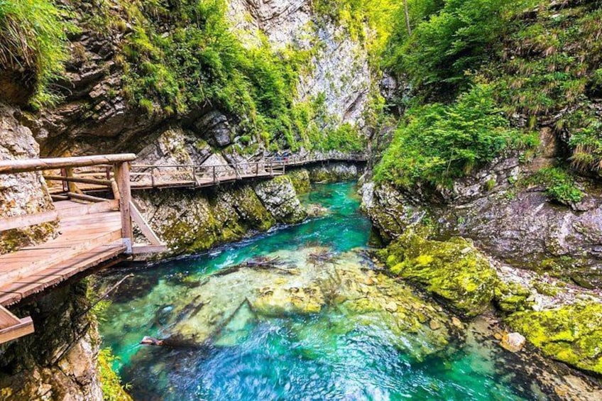 A wooden bridge over a clear blue stream surrounded by rocky cliffs and greenery. 2