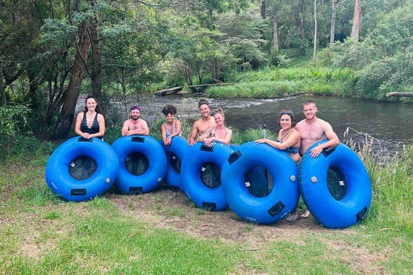 A group of people sitting on blue inflatable rings by a river in a forested area. 5