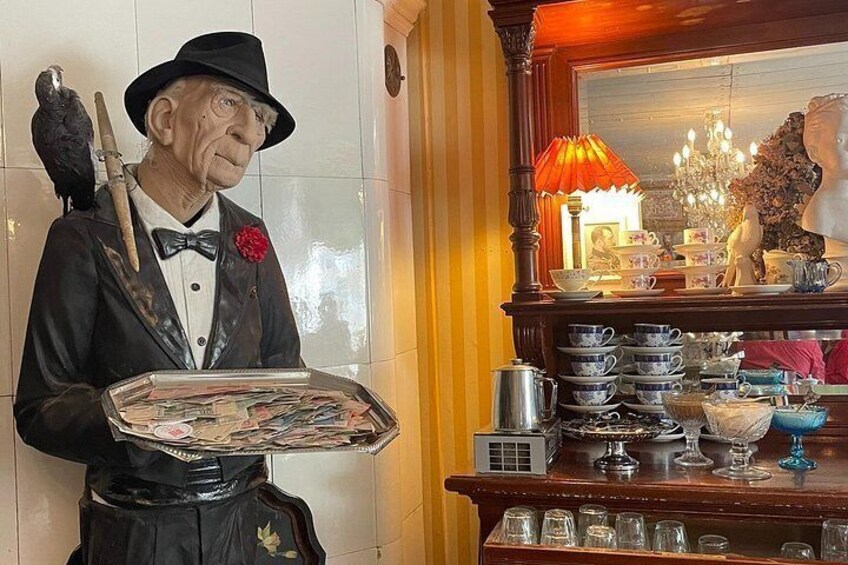 Una estatua de un hombre trajeado sosteniendo una bandeja, con un pájaro posado en su hombro y un juego de té sobre una mesa detrás. 3
