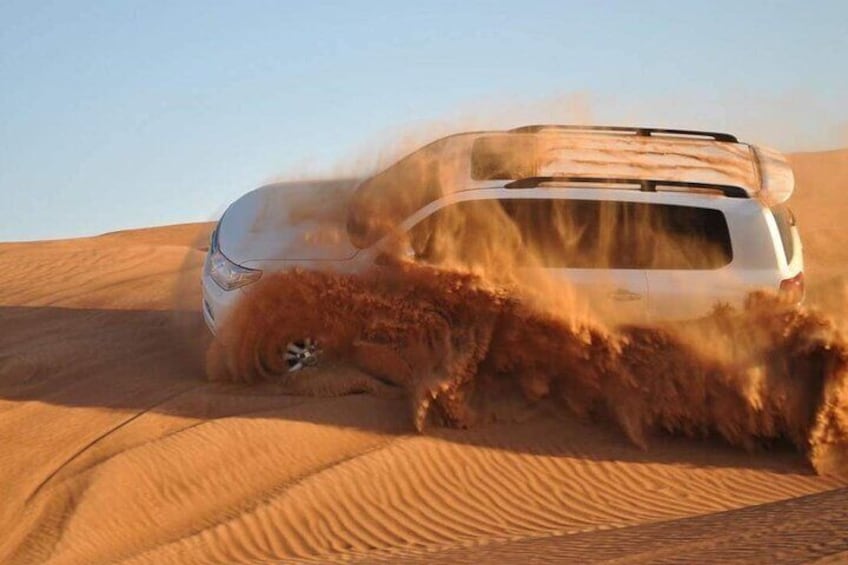 A white SUV driving through a desert with sand being kicked up around it. 1