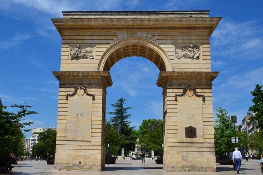 Une grande arche en pierre ornée de sculptures en relief, entourée d’arbres et de bâtiments. 1