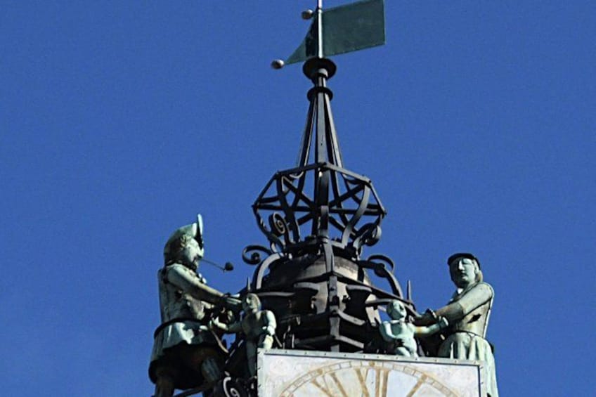 Une statue représentant deux personnages, posée sur un piédestal, et surmontée d’une girouette et d’une boussole. 2