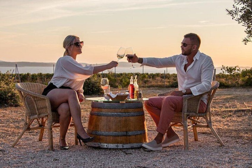 Two people toasting with wine glasses at a round wooden table with bottles, outdoors with a view of the sea. 1