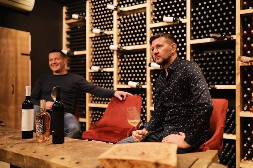 Two men seated at a table with wine bottles and glasses, in a room with wine racks in the background. 4