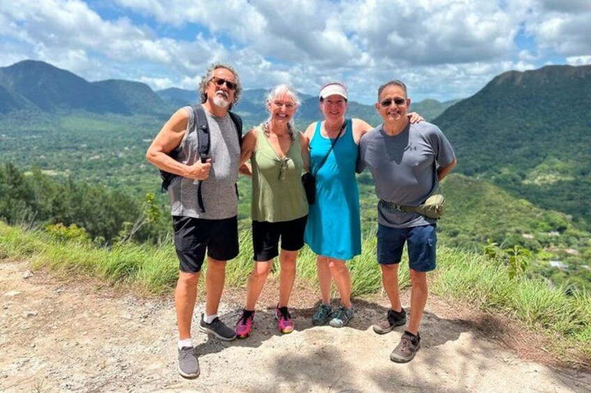 Four people standing on a mountain trail with a scenic view of mountains and greenery in the background. 1