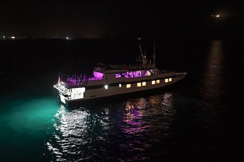 A large, illuminated boat with purple lighting on its deck, floating on calm water at night. 5