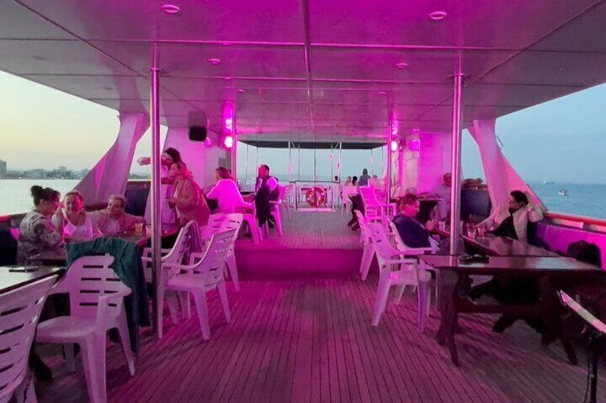 A boat deck with people dining and socializing, wooden flooring, and a view of the water. 3