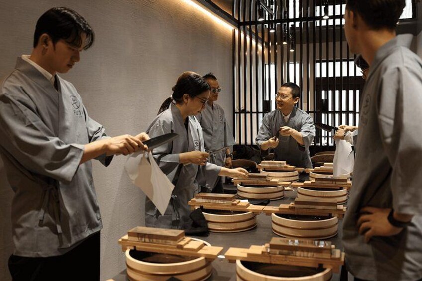 A group of people in a kitchen setting, some wearing aprons, preparing food with wooden bowls and utensils. 5