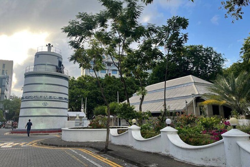 A cylindrical monument with inscriptions, a building with a sloped roof, and a landscaped area with trees and flowers. 1