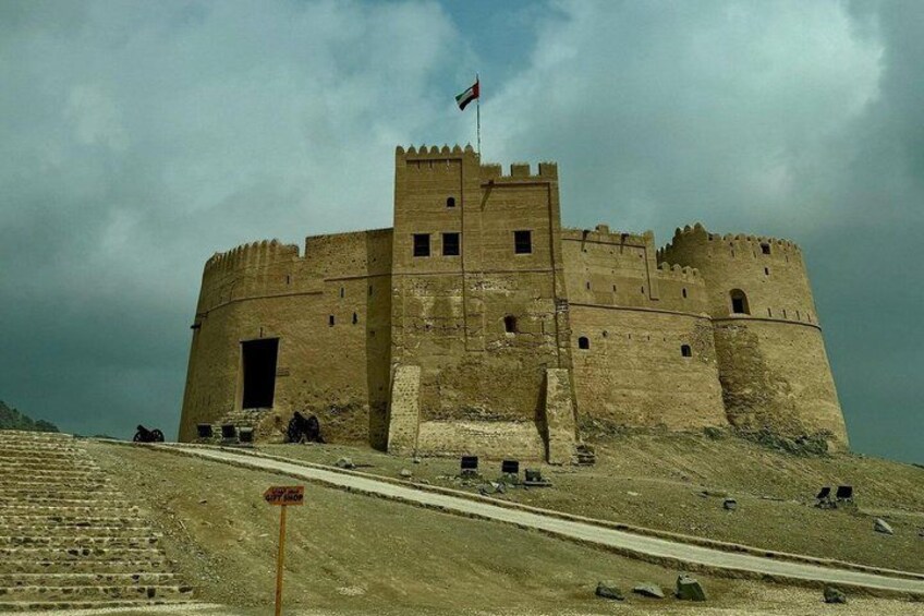 A large, historic stone castle with a flag on top, situated on a hill with a dirt road leading up to it. 5