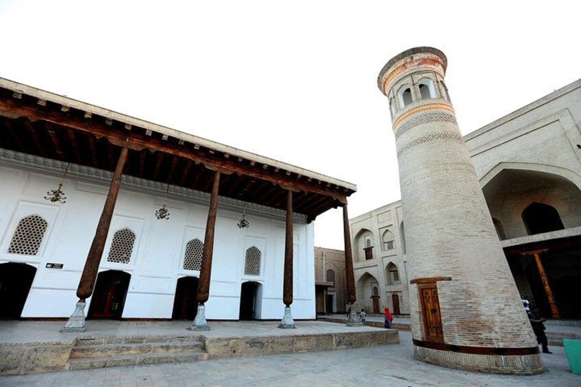 A courtyard with a tall, cylindrical tower, a white building with arched doorways, and a wooden roof structure. 4