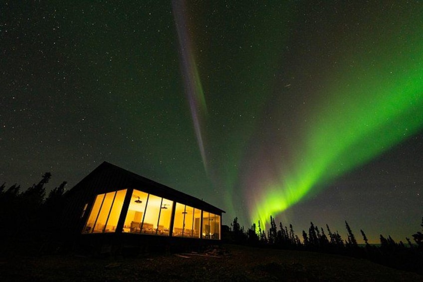 A building with large windows illuminated at night, under a night sky with green aurora borealis. 2
