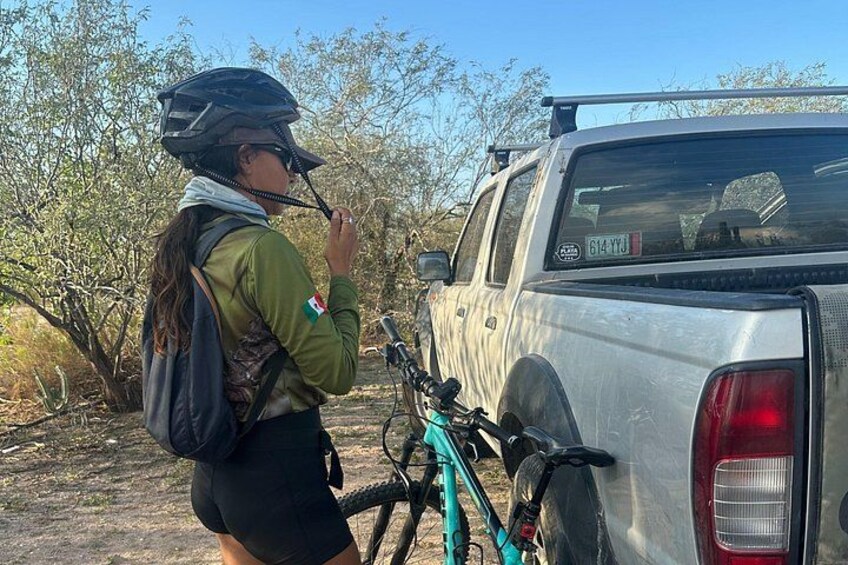 A person with a helmet and backpack standing next to a silver pickup truck with a bicycle mounted on the back. 5
