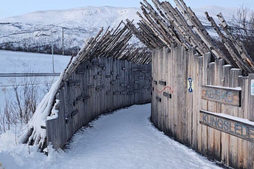 En trækonstruktion med et buet tag, snedækket jord og et skilt der står 'ABISKO KUNGSLEID'. 2