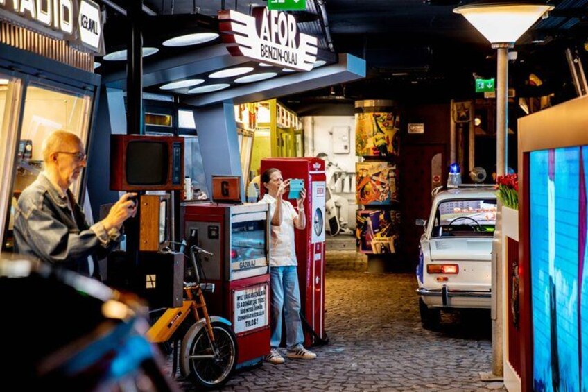 A vintage-themed indoor setting with retro arcade machines, a vintage car, and a person using a vintage telephone. 1