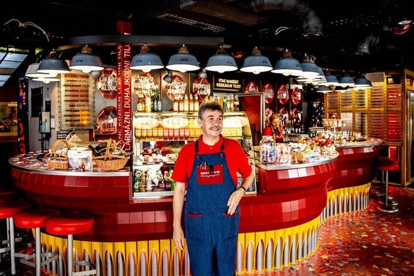A person standing in front of a brightly lit food stall with a red and yellow color scheme, various food items displayed, and a patterned floor. 4