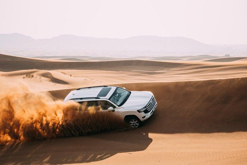 A white SUV driving through a desert with sand dunes and mountains in the background. 1