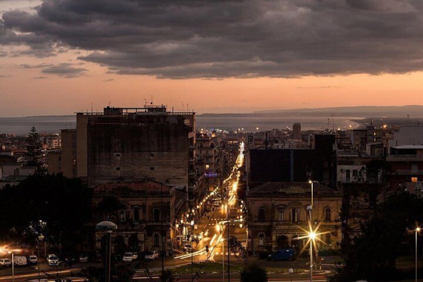A cityscape at dusk with a prominent street lined with buildings and streetlights. 2