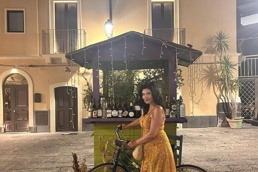 A woman in a yellow dress standing next to a bicycle, with a kiosk in the background. 3