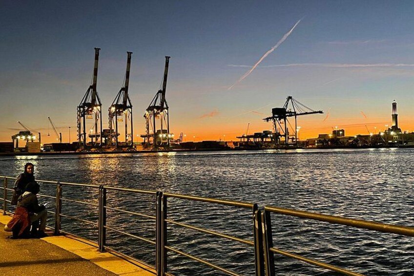 Un muelle con grúas industriales y una persona sentada al atardecer, frente al agua. 3