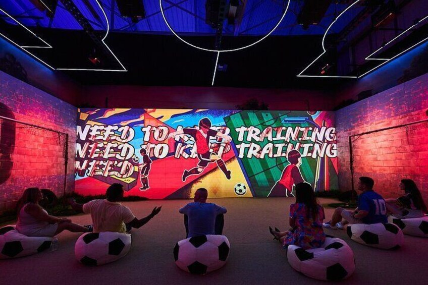 A group of people sitting on soccer balls facing a colorful mural with the text "NEED TO TRAINING NEED TO TRAINING". 4