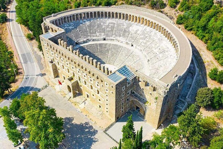An ancient amphitheater with a semi-circular structure, surrounded by greenery and a paved road. 5