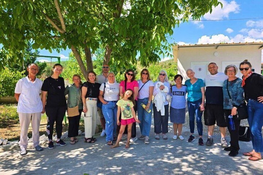 A group of people standing outdoors under a tree, with a building in the background. 1