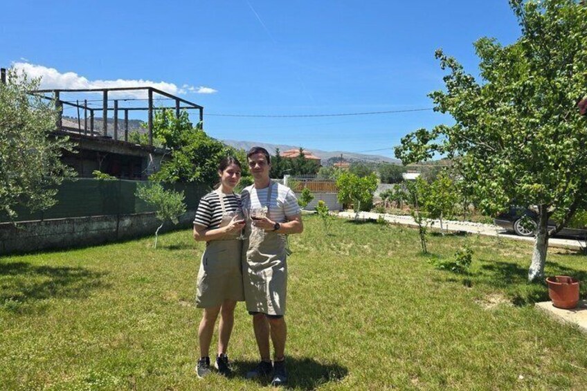Two people standing in a grassy area with a tree and a building in the background. 3