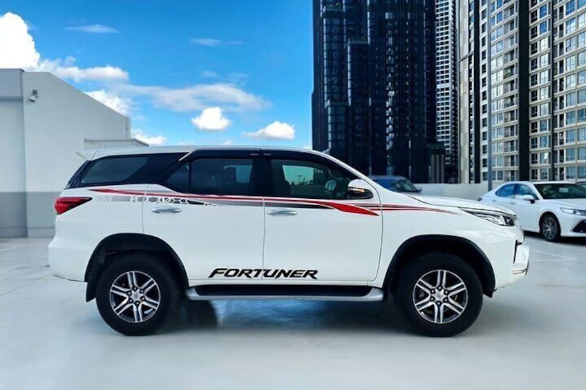 En hvid SUV med 'FORTUNER' branding parkeret i et bymiljø med moderne bygninger i baggrunden. 5