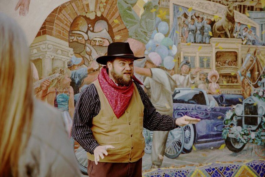 A person in a hat and vest pointing towards a mural with a car and people. 3