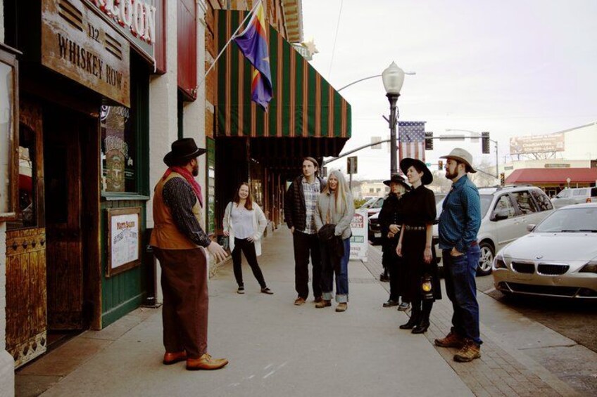 A group of people standing on a sidewalk in front of a building with a sign that reads 'WHISKEY ROW.' 1
