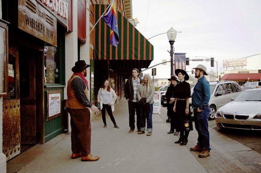 A group of people standing on a sidewalk in front of a building with a sign that reads 'WHISKEY ROW.' 1