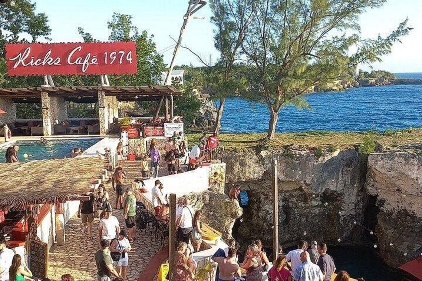 A crowded outdoor café with a pool, people sitting and standing, and a rocky shoreline. 3