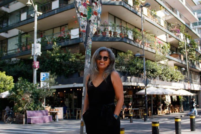 A person standing in front of a building with balconies, a street with bollards, and outdoor seating. 4