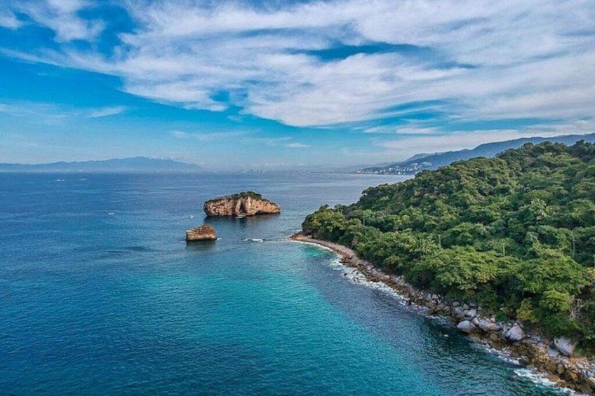 A coastal landscape with a rocky outcrop surrounded by clear blue waters and a densely vegetated hill. 1