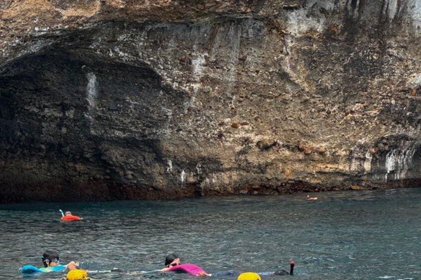 A rocky cliff by the sea with a person swimming and a red boat in the water. 5