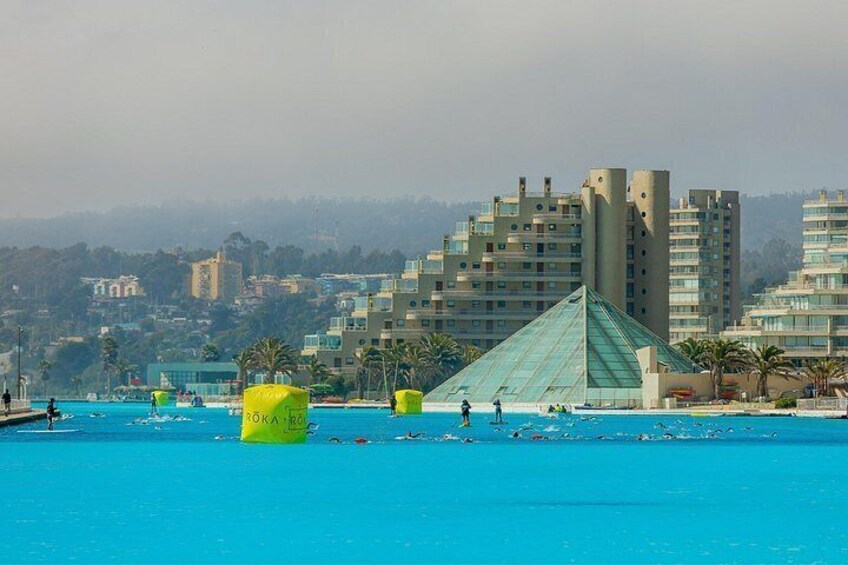 Une grande piscine où se baignent des gens, un bâtiment en forme de pyramide et, au loin, des immeubles de grande hauteur. 5