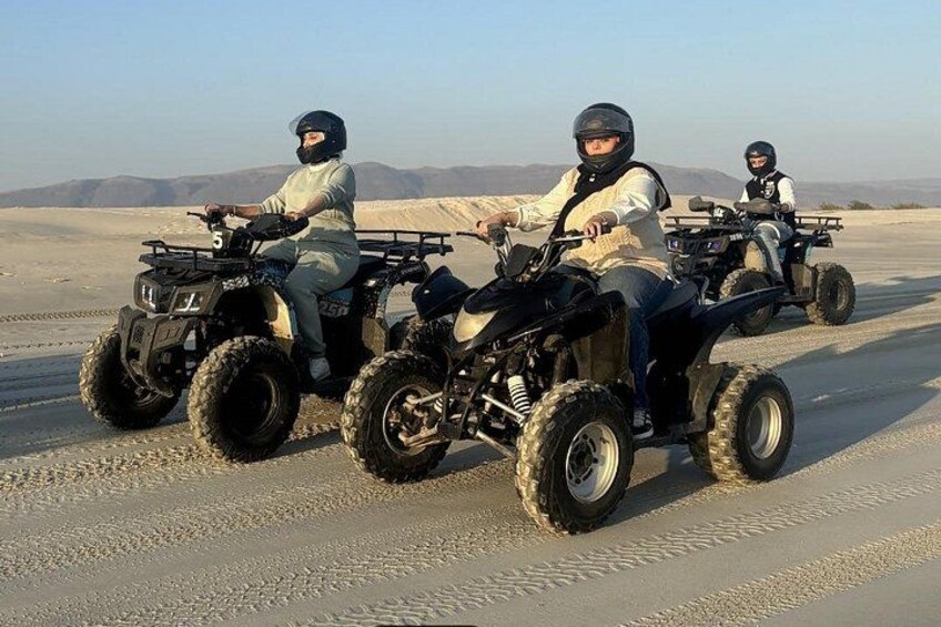 Three people riding ATVs on a desert road with sand dunes in the background. 3