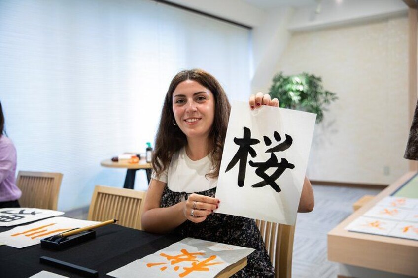 Een persoon houdt een wit vel papier met zwarte Japanse karakters vast, zittend aan een tafel met een donker oppervlak en een pen. 3