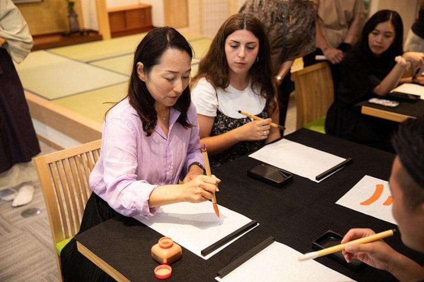 A person writing on a piece of paper with a pen, a smartphone, and a small orange object on the table. 4