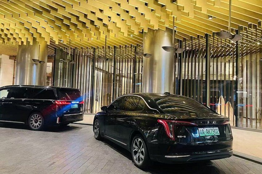 A black car parked in front of a modern building with a patterned ceiling and vertical columns. 1